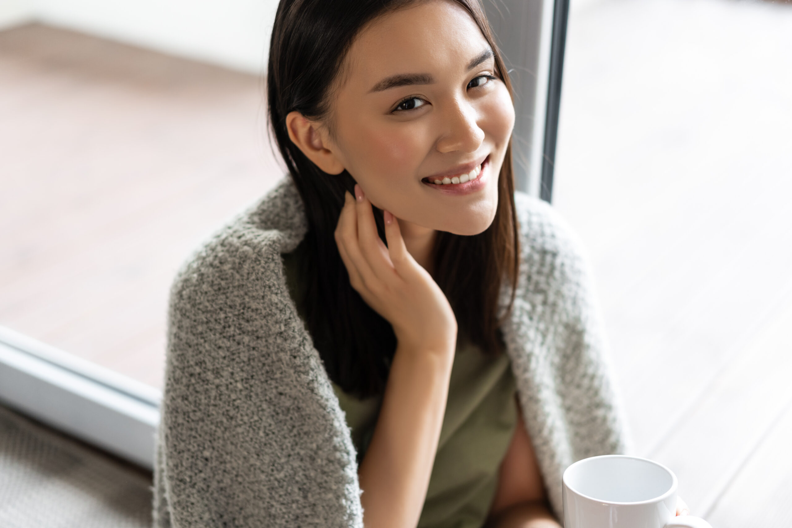 Portrait of beautiful asian woman wrapped in blanket, drinking coffee and smiling, relaxing at home on weekend, sitting near window and enjoying the view